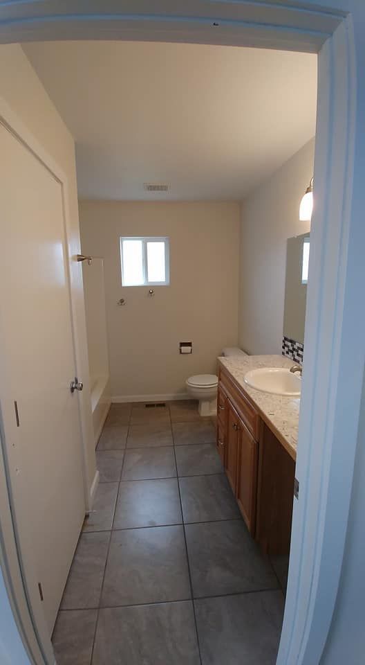 Narrow bathroom with tiled floor, wood vanity and sink on the right, toilet at the back, and a small window.