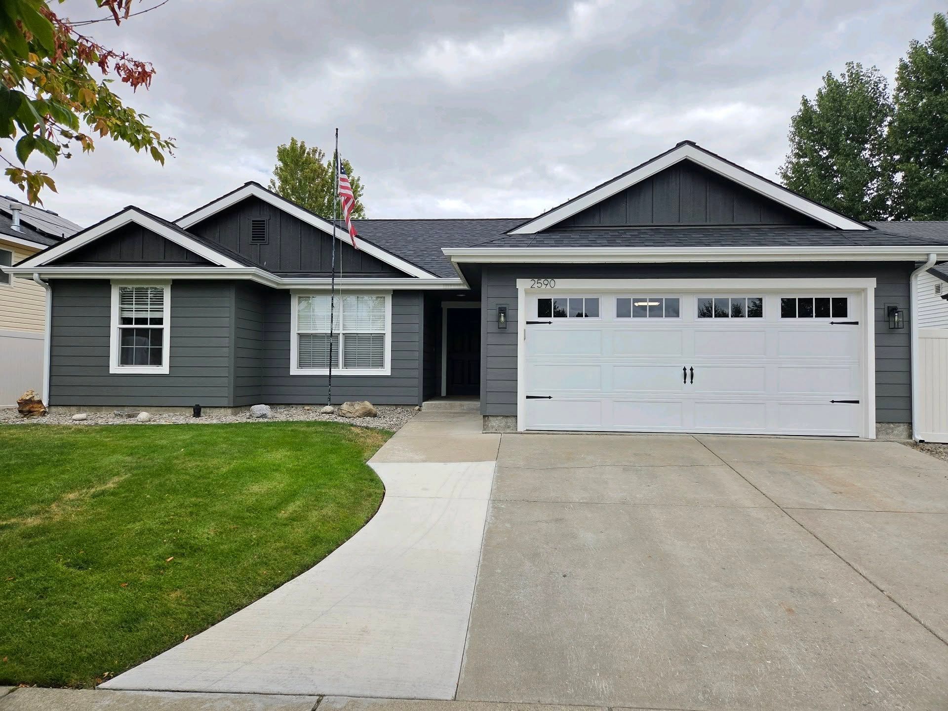 Gray single-story house with white garage, front lawn, and curved concrete walkway under an overcast sky
