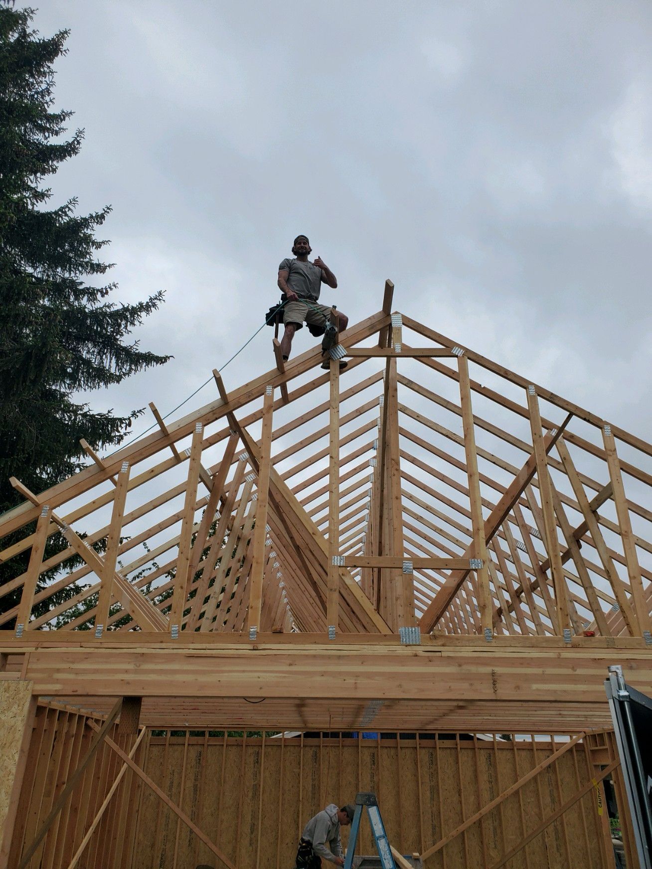 Worker standing on a house roof frame under construction, with wooden trusses and a cloudy sky behind.