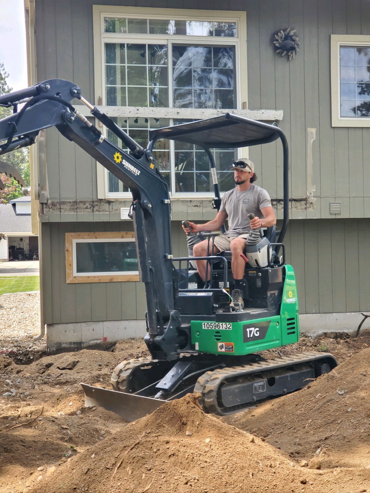 Mini excavator digging dirt beside a house, with a worker operating it.