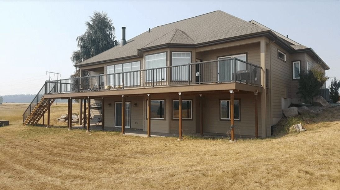 Two-story beige house with a wraparound deck and dry grass yard under a clear sky