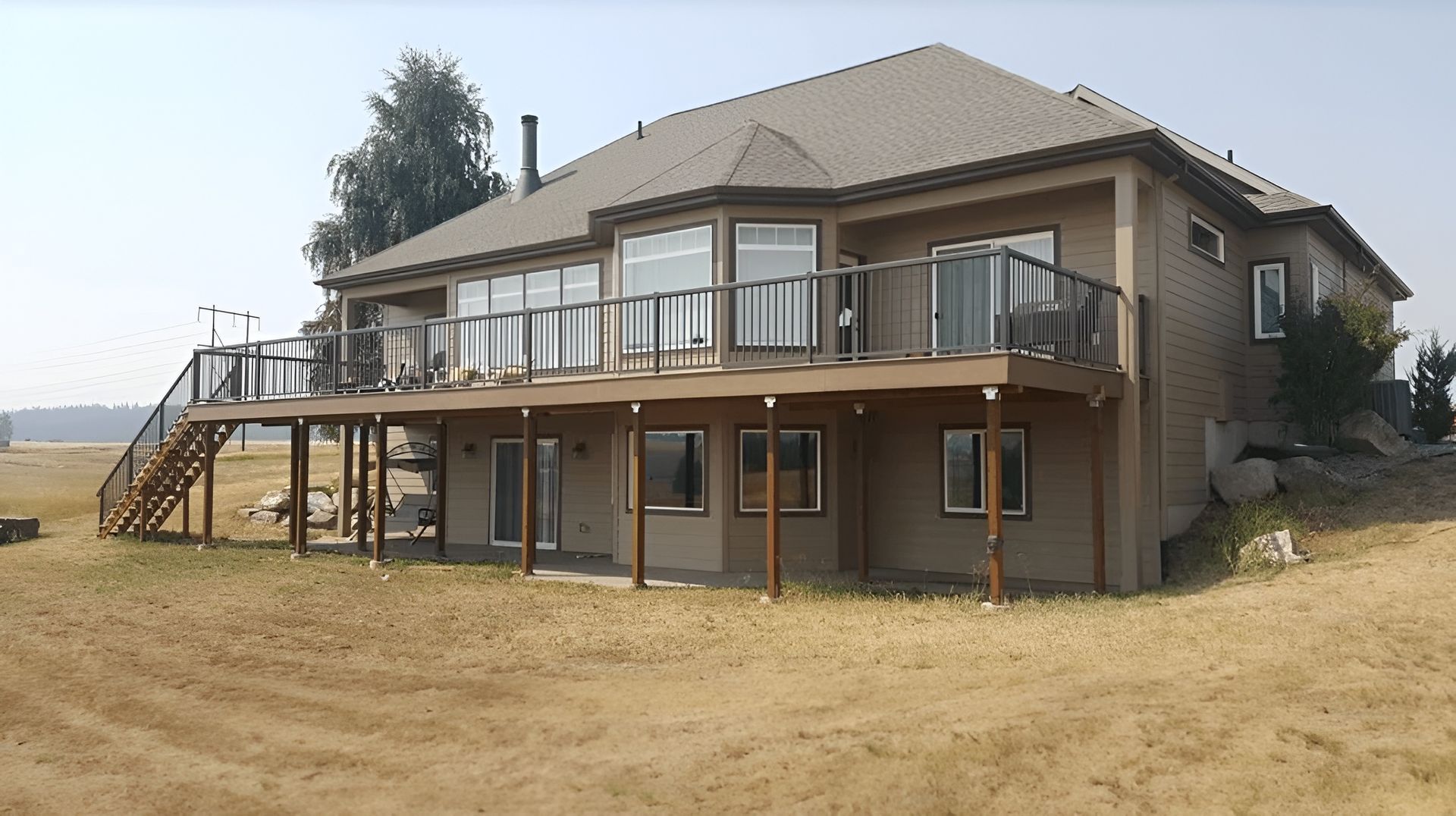 Two-story beige house with a wraparound deck on a dry grassy lot under a clear sky