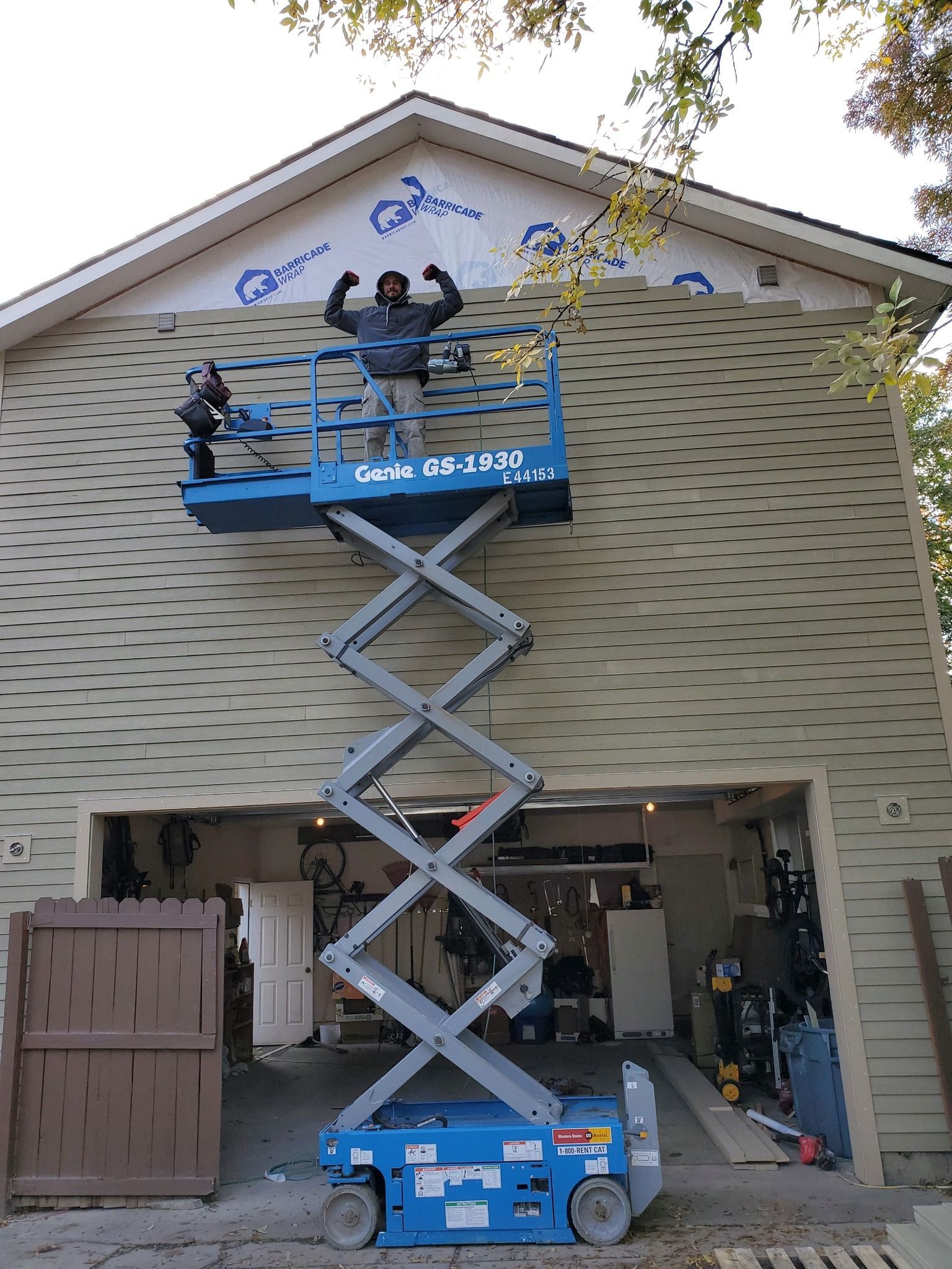 Worker in blue lift inspecting house roof with scissor lift at open garage.
