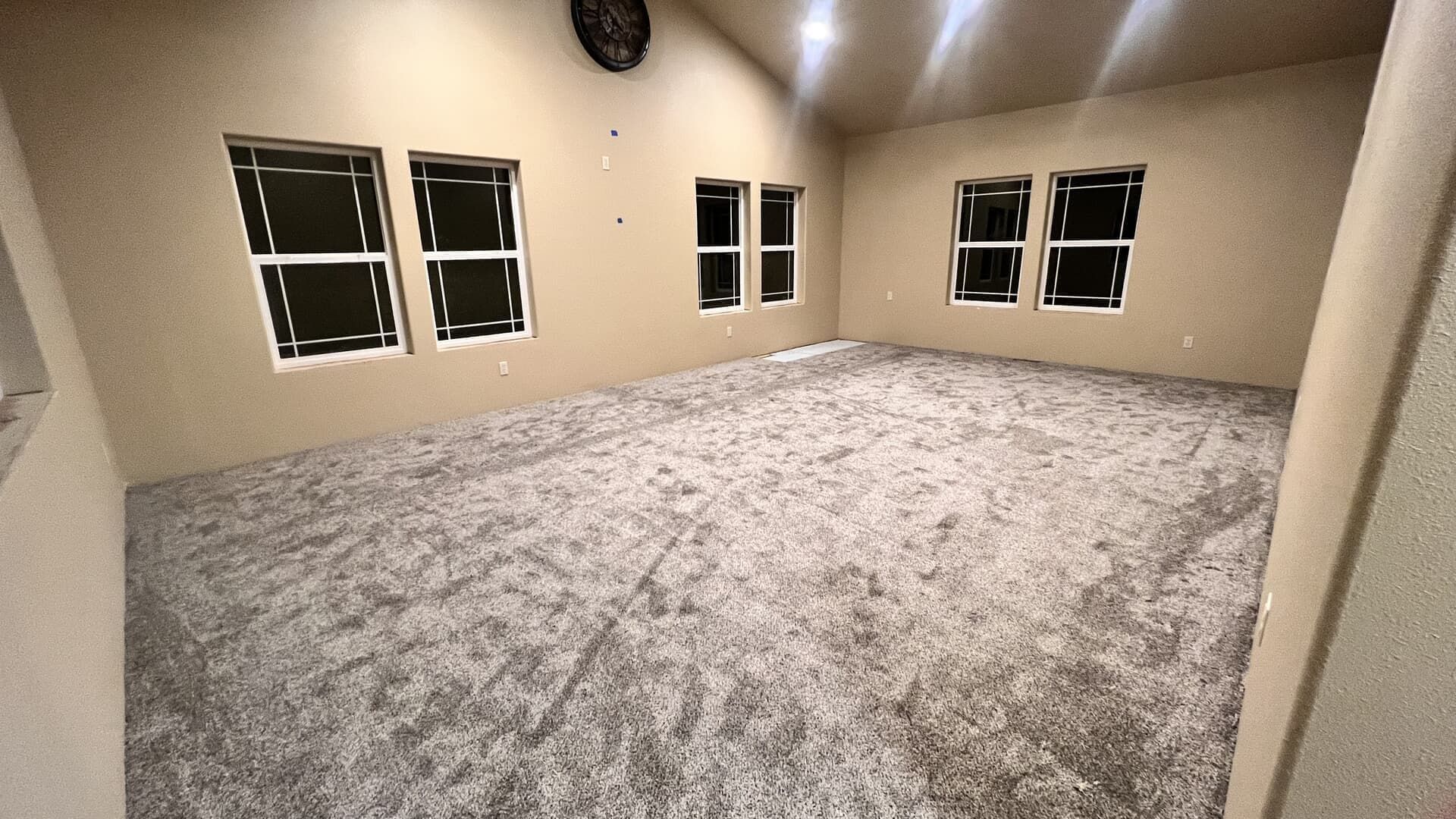 Empty carpeted room with beige walls, multiple windows, and recessed ceiling lights