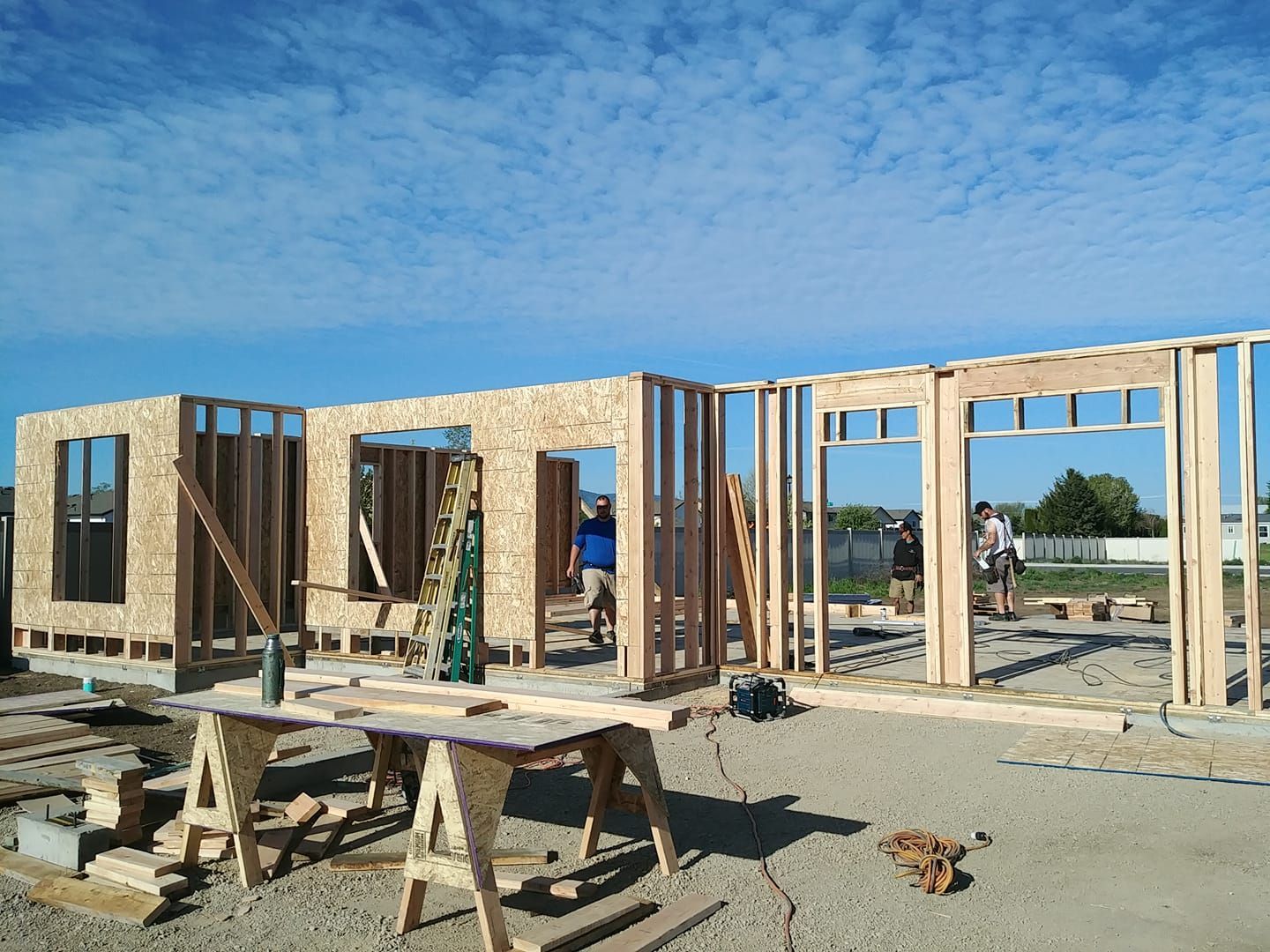 Wood-frame house under construction with workers, blue sky, and scaffolding on a concrete slab