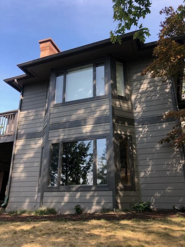 Two-story gray house with large windows, dark roof, and a brick chimney in a wooded yard