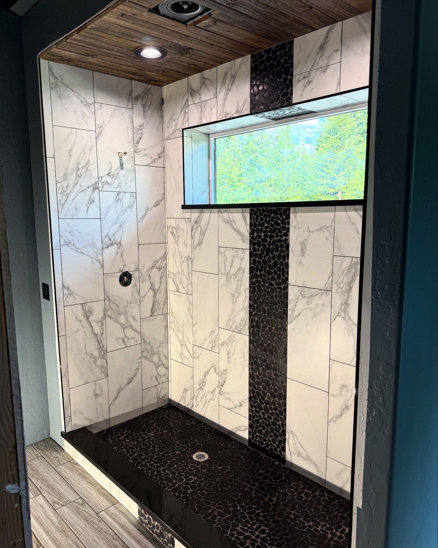 Modern shower stall with marble-pattern walls, black floor, wood ceiling, and a wall-mounted screen.