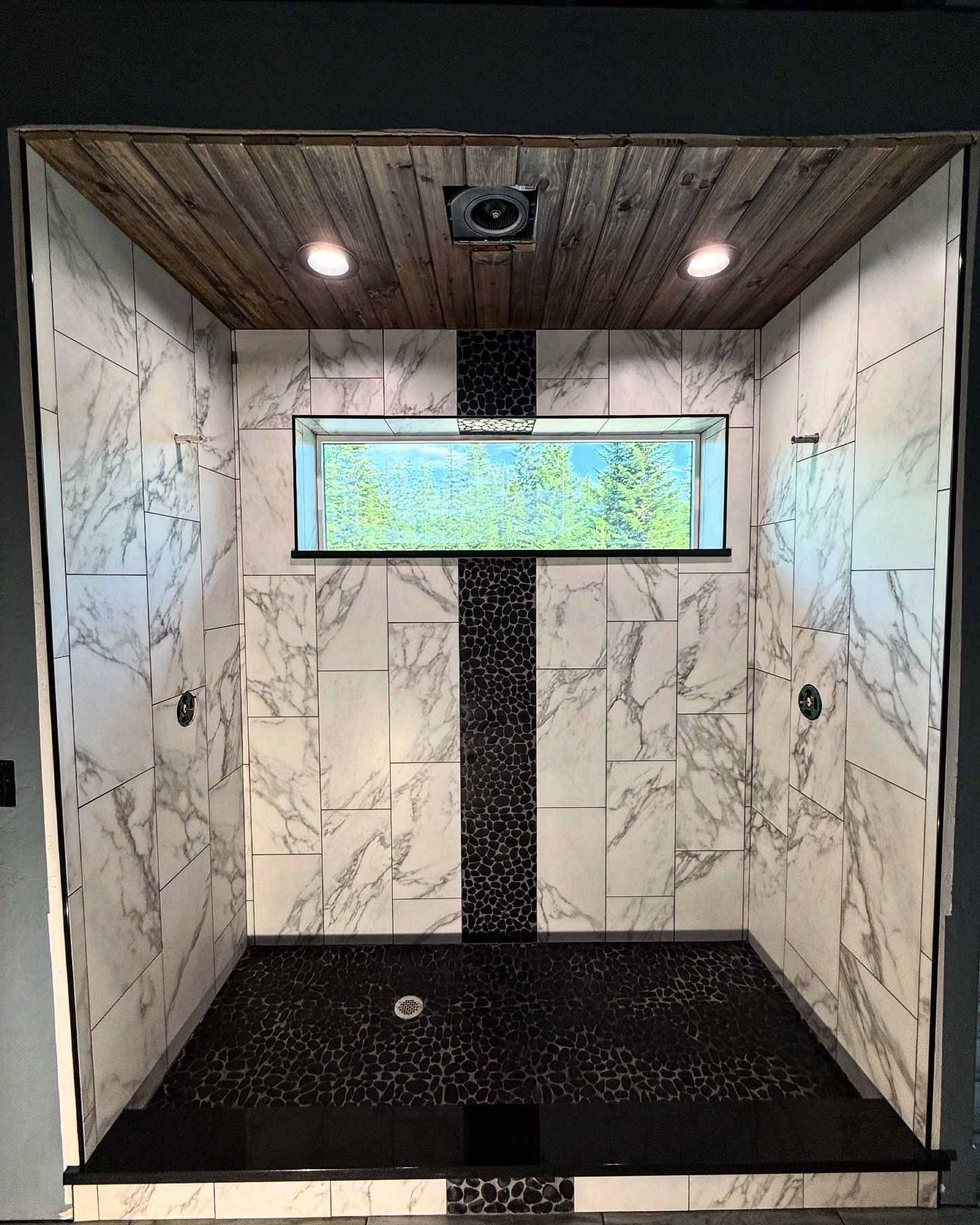 Empty marble-tiled shower with black floor, recessed lights, and a narrow window.