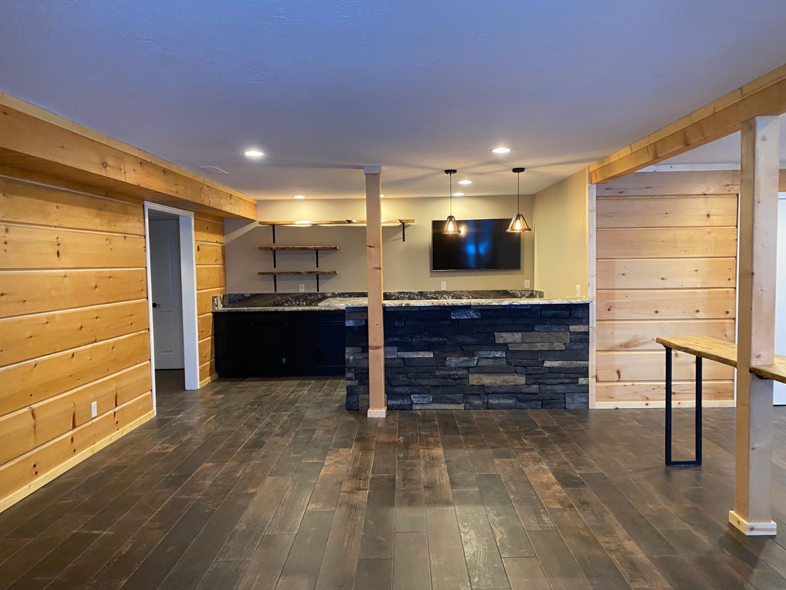 Empty basement rec room with wood-paneled walls, dark floor, stone bar, and TV mounted at the back.