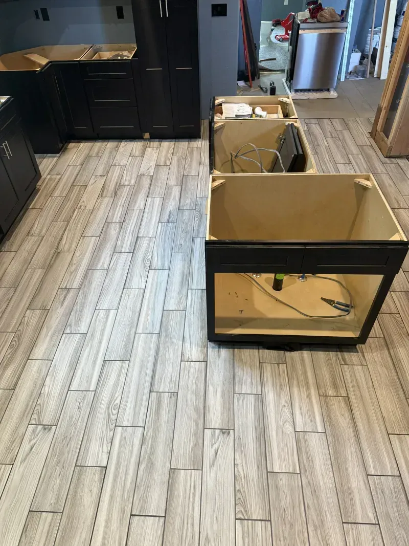 Two open cardboard boxes on a wood-look kitchen floor beside black cabinets and a hallway.