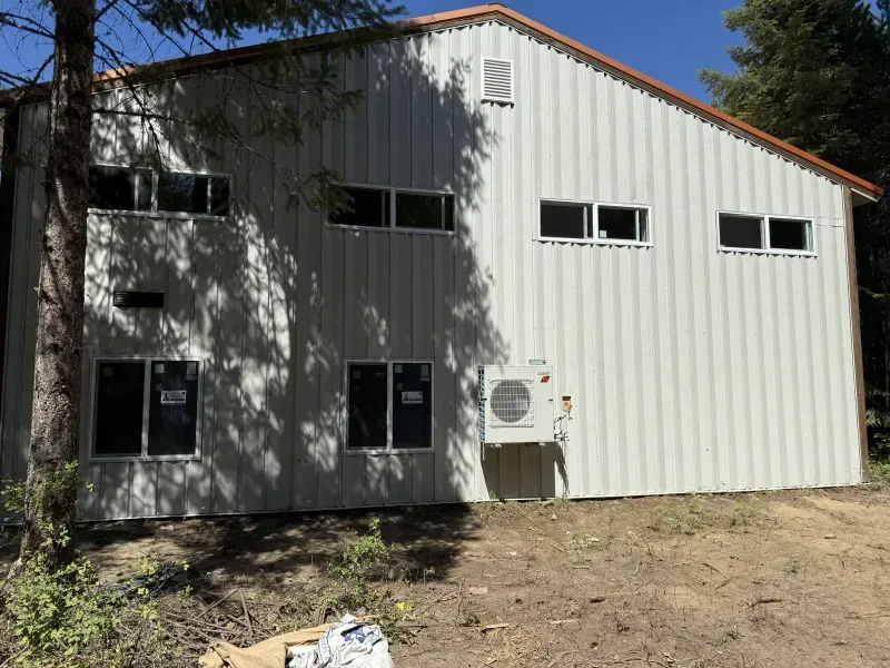White metal building with several windows and doors, air-conditioning unit, and dirt yard in front