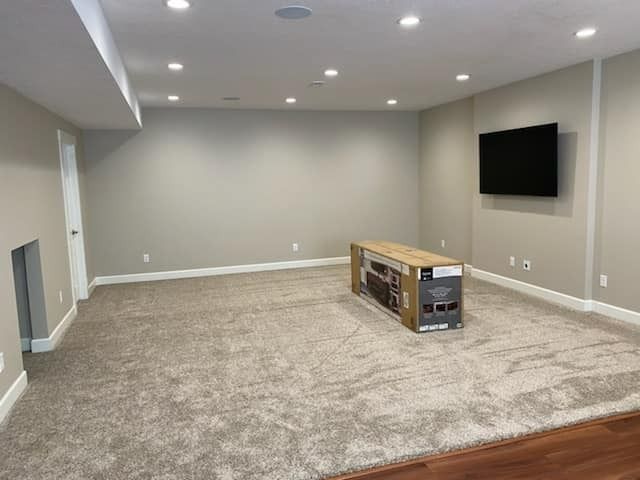 Empty finished basement with beige carpet, gray walls, recessed lights, and a wall-mounted TV.