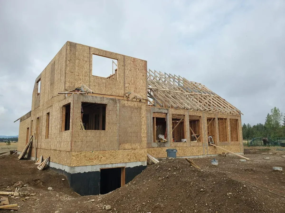 Unfinished two-story wooden house under construction on a dirt lot, with exposed framing and roof trusses.