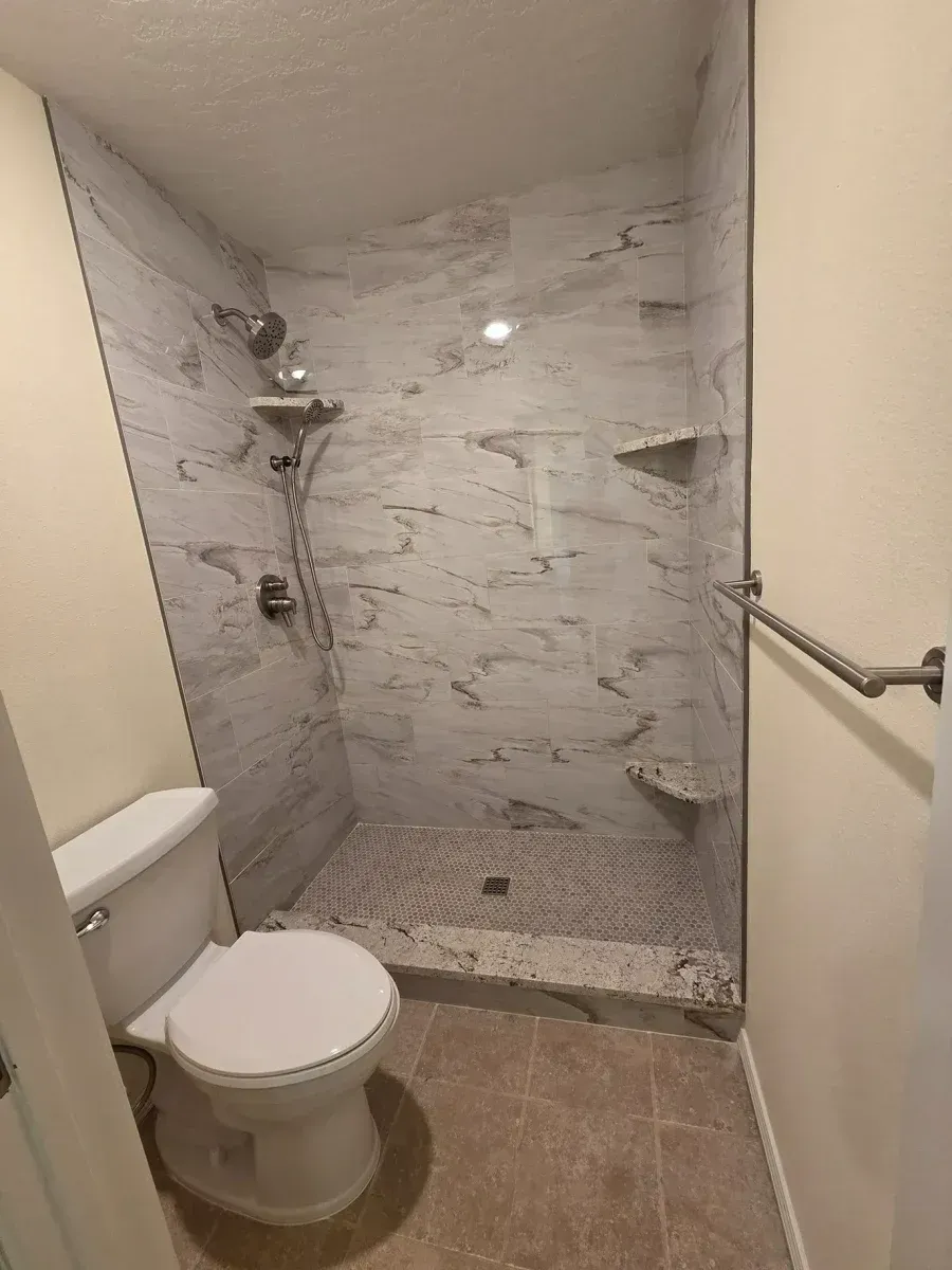 Compact bathroom with walk-in shower, marble-look tile, toilet, and grab bar.