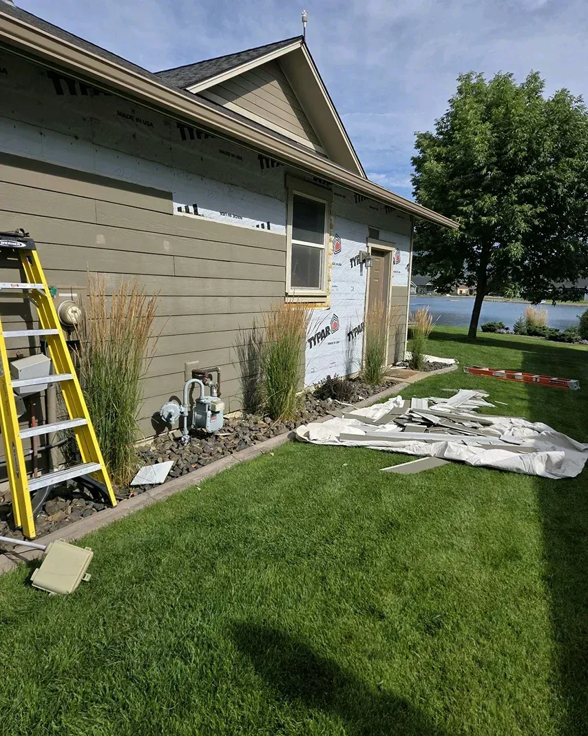 House exterior with yellow ladder, white tarp on grass, and a lake visible in the background.