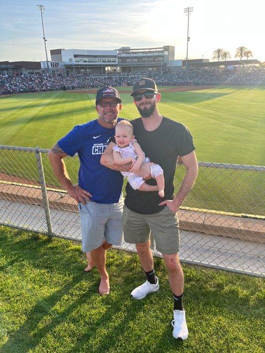 Two adults and a baby smiling on a baseball field, with a crowd in the stands behind them.