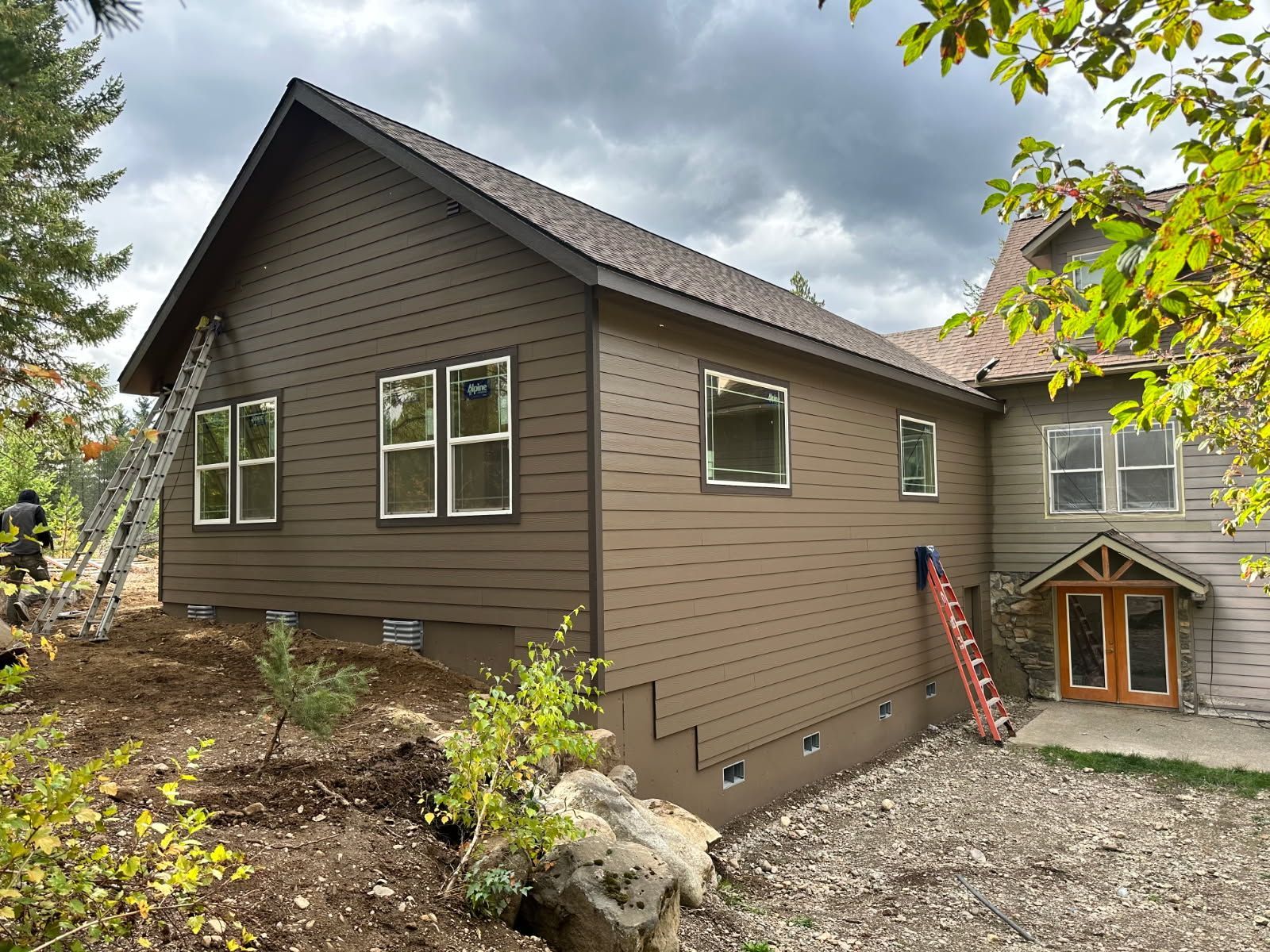 Brown house exterior with windows, gravel yard, and a small entryway under cloudy skies