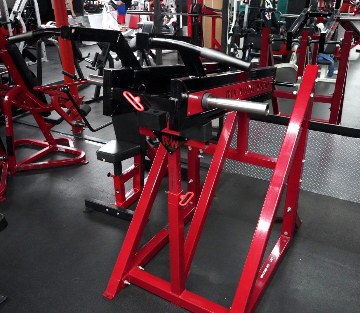 Red and black gym exercise machines in a weight room with rubber flooring.