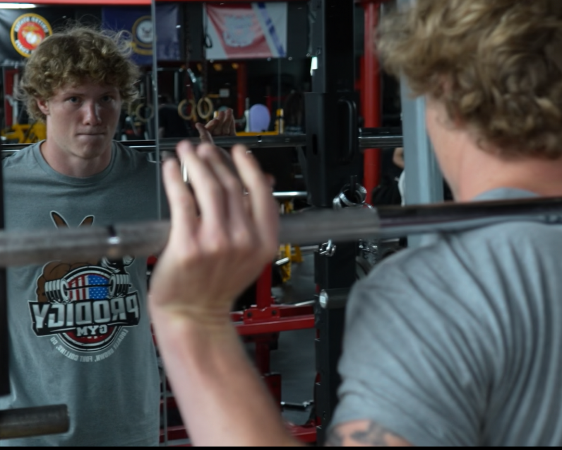 Man lifting a barbell in a gym, seen in a mirror with weight racks behind him.