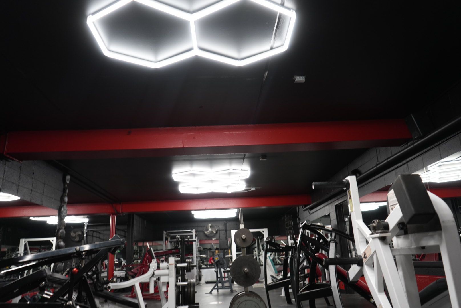 Dim gym interior with red accents, mirrored walls, weight machines, and bright geometric ceiling lights