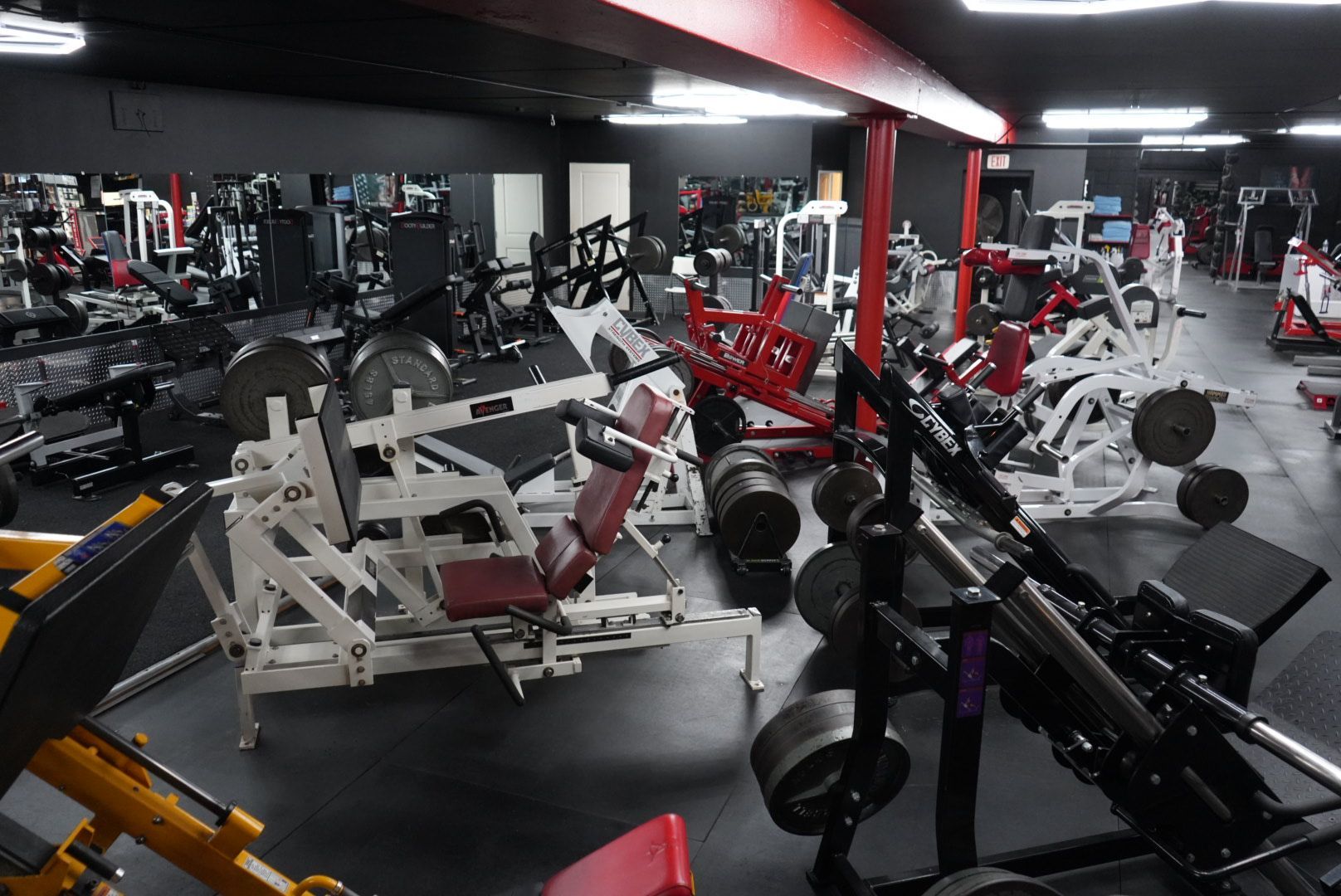 Gym interior with weight machines and benches, mostly white and red equipment under bright overhead lighting