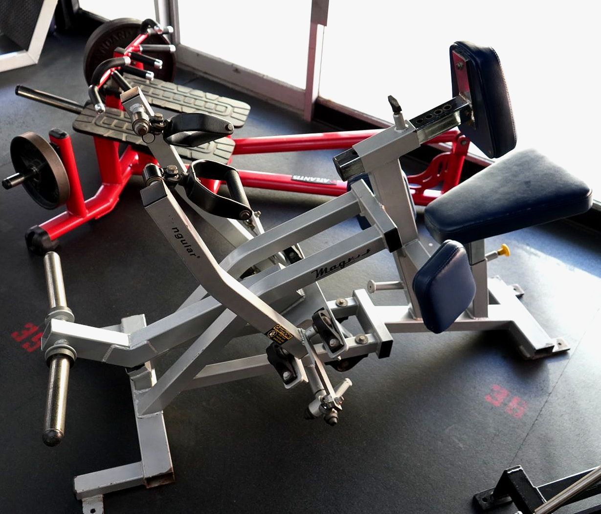 A red and silver leg press machine with padded seat and footplate in a gym.