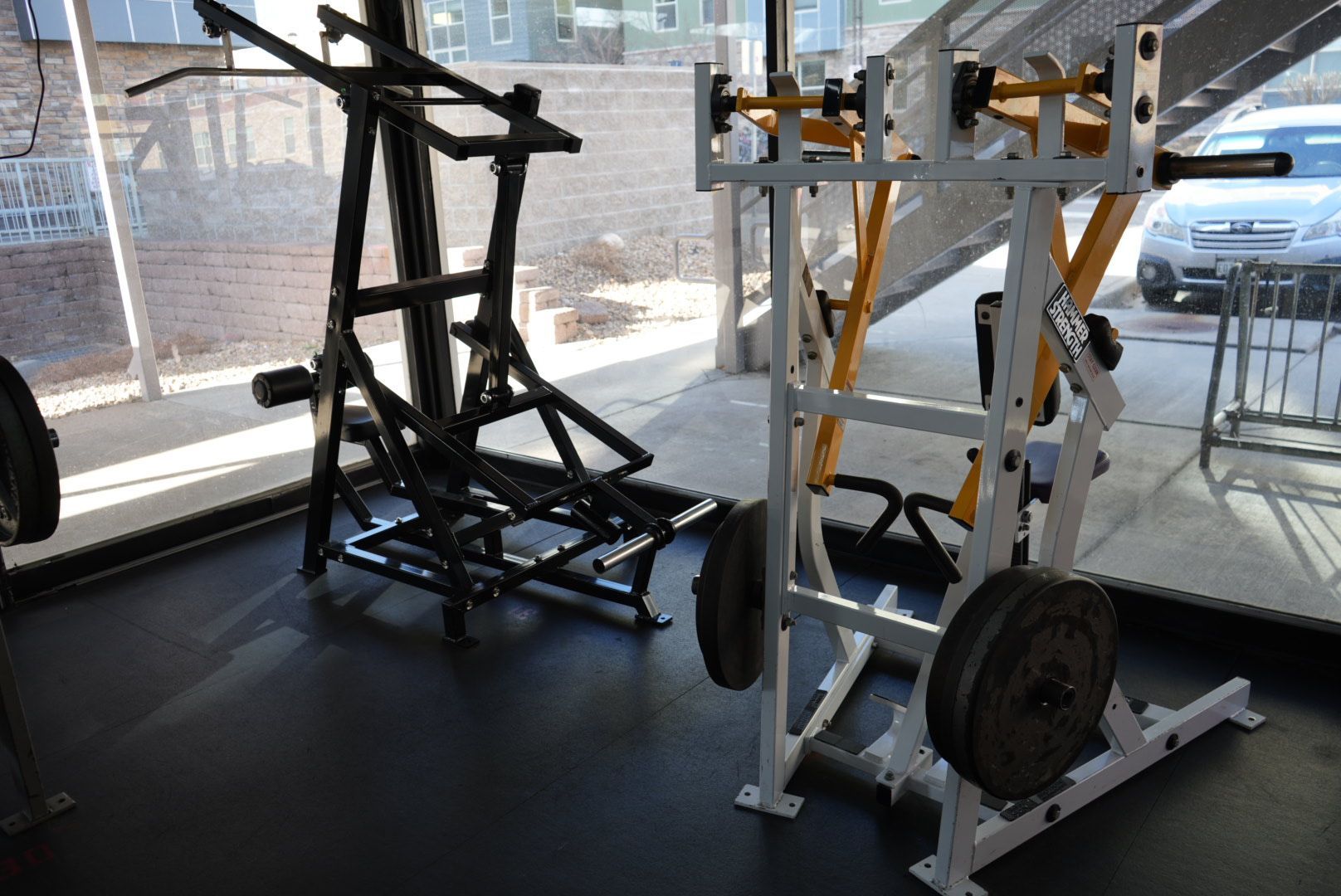 Two black metal exercise frames with wheel bases in a gym by large windows