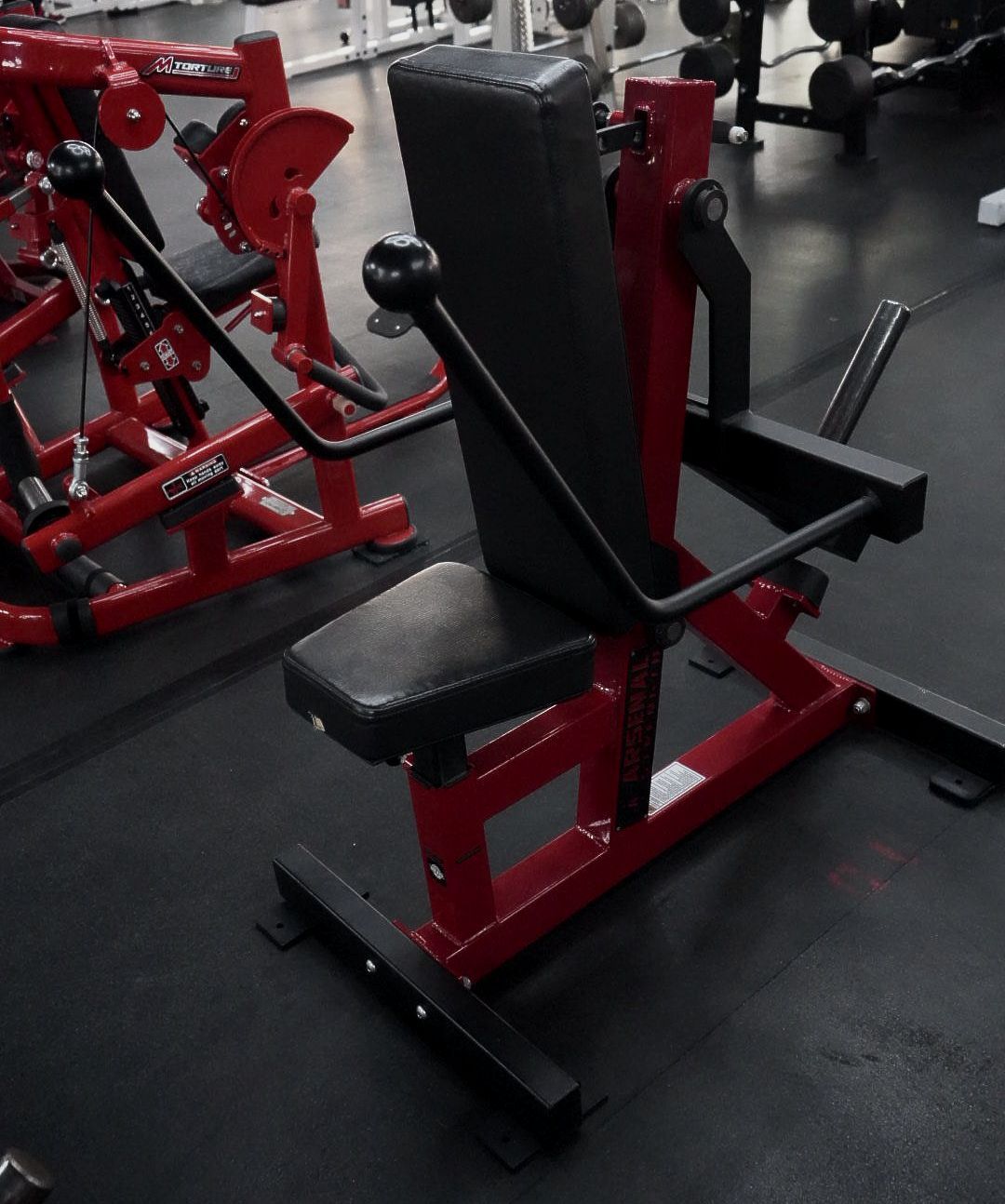 Red and black gym exercise machine with padded seat and backrest in a fitness room.