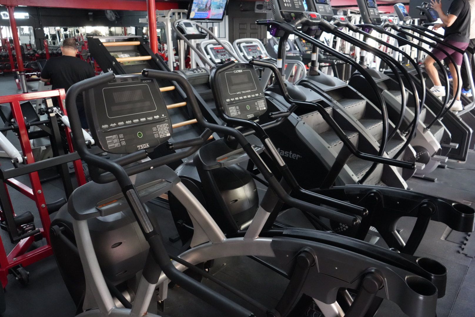 Rows of black and red ellipticals in a gym, with a person exercising in the background.