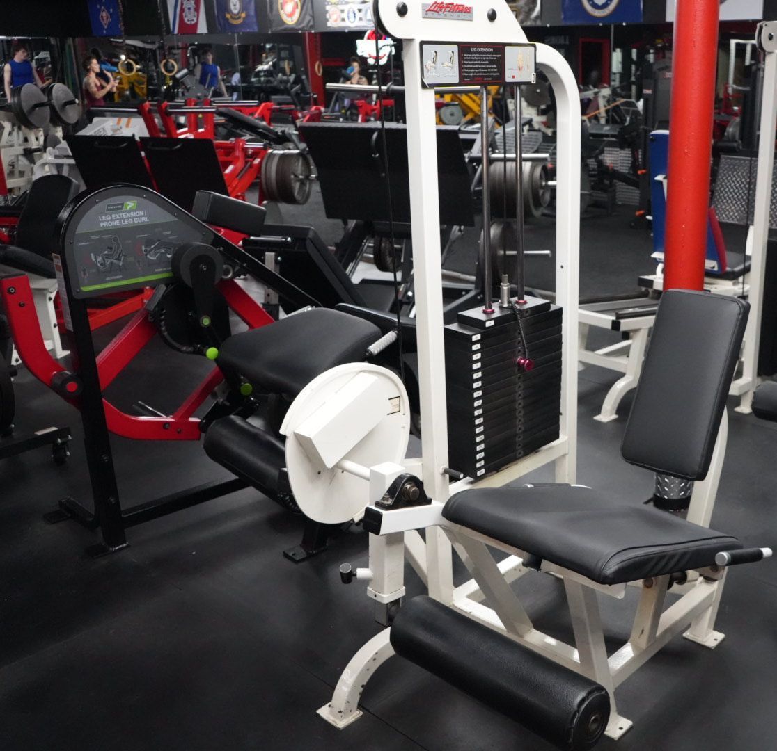 Gym machine with black padding and white frame in a fitness center, with red exercise equipment in the background