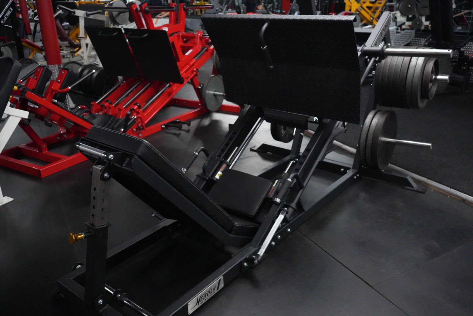 Red and black weight machine in a gym, with angled padded seats and metal frames.