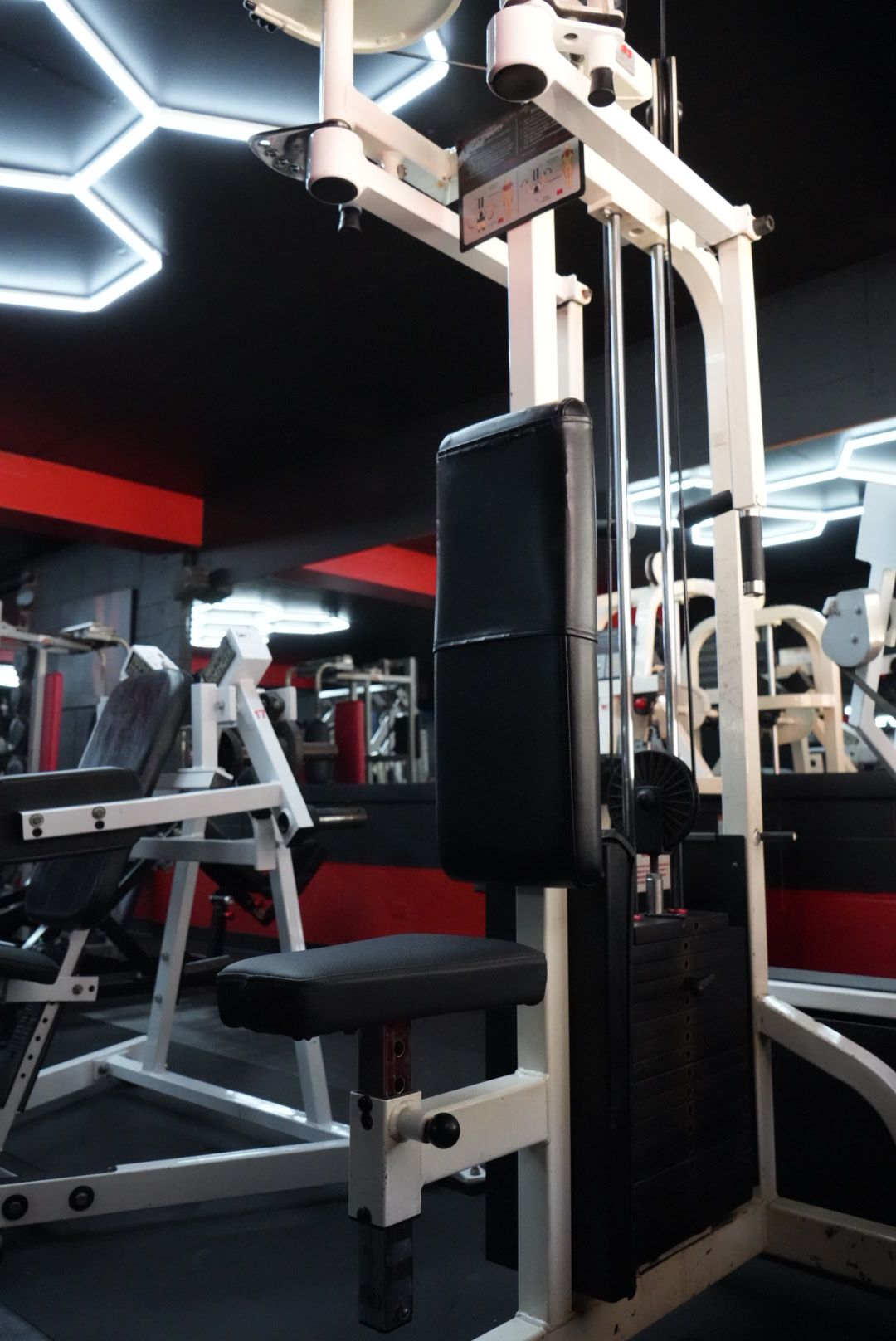 Cable weight machine in a gym, with black padding and white frame under bright geometric ceiling lights