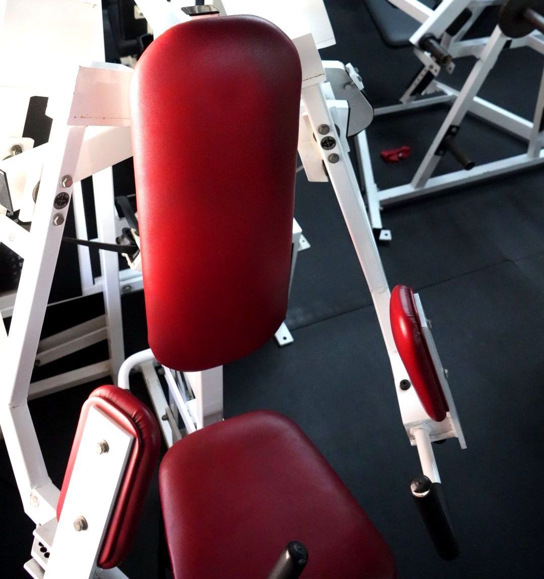 Red padded weight machine seat and backrest in a gym, with white metal frame and black flooring.