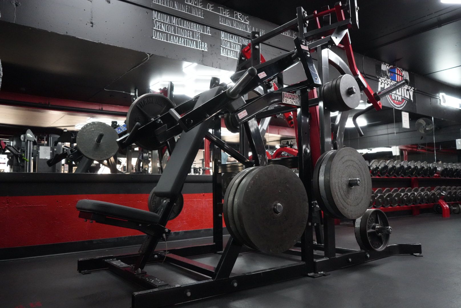 Two black gym machines in a red-and-black weight room with mirrors and plate-loaded weights