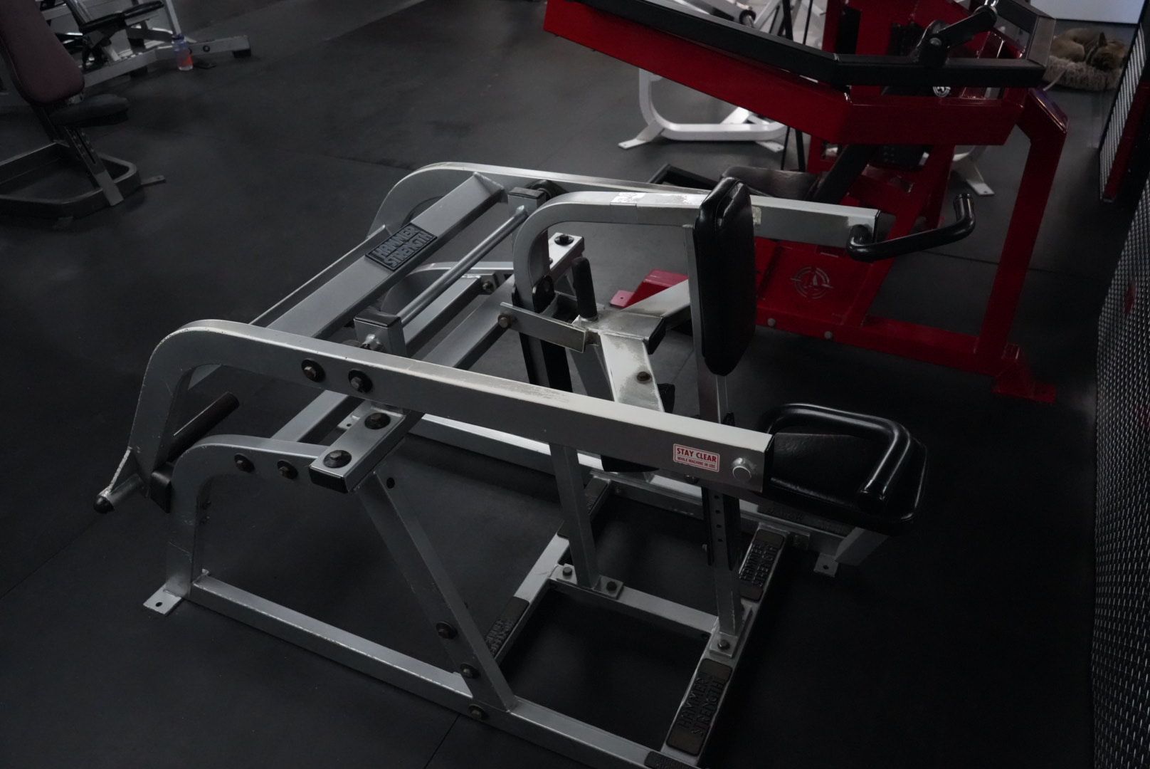 Gym leg press machine in a dark workout room, with silver frame and black seat.