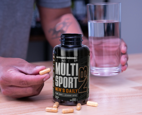 Hand holding a capsule beside a bottle of multivitamin supplements and a glass of water on a table