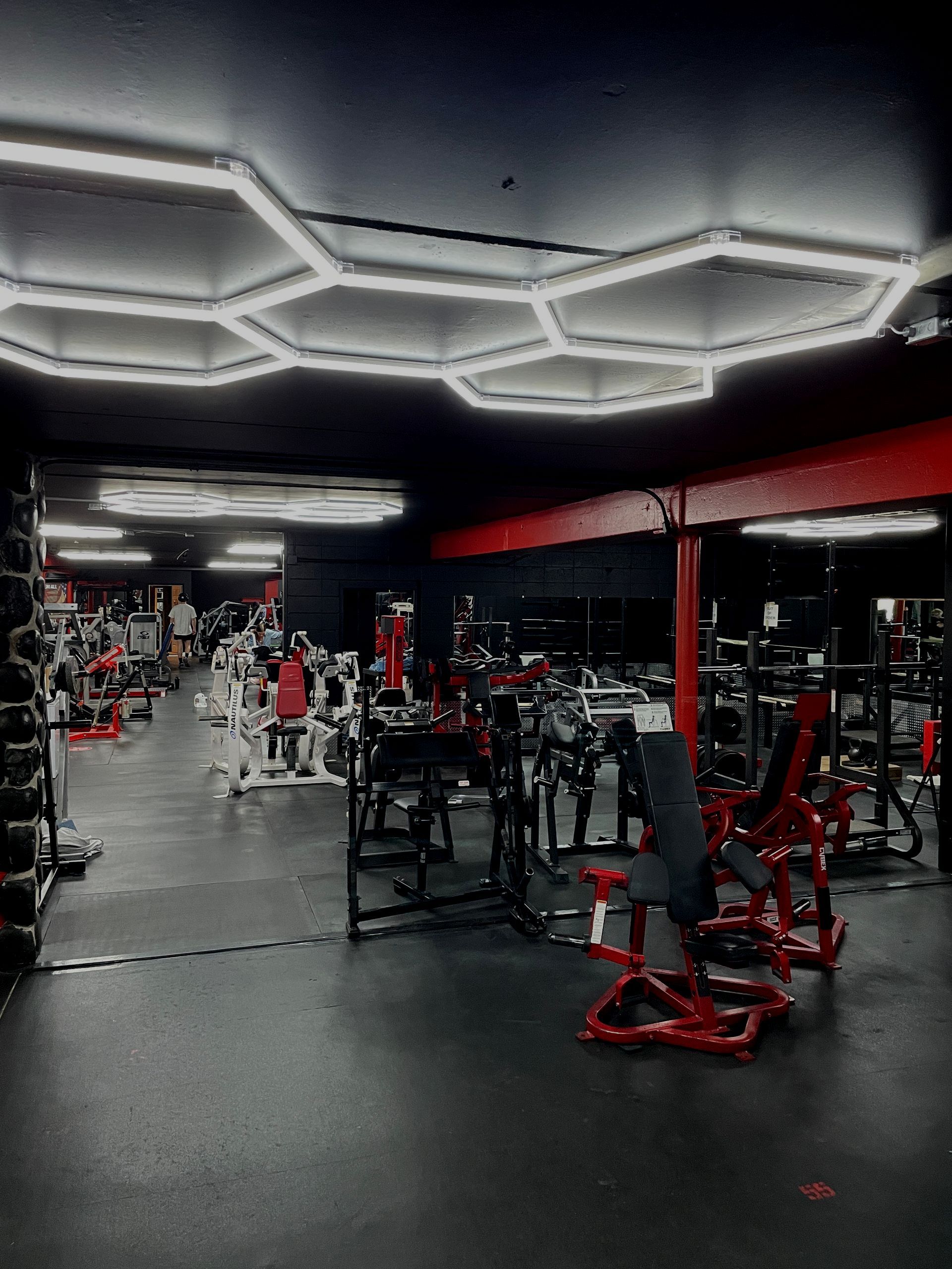 Empty gym with red equipment, black flooring, and bright hexagonal ceiling lights.