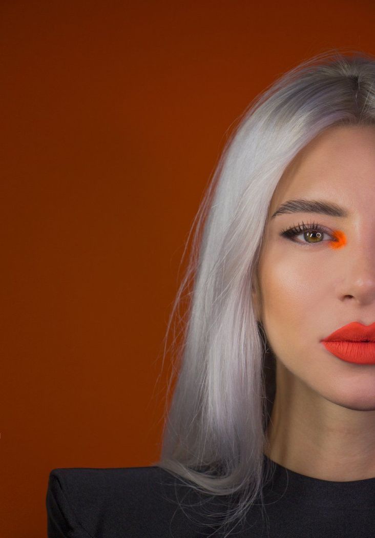 Primo piano di una donna con capelli argentati e trucco arancione su uno sfondo arancione.