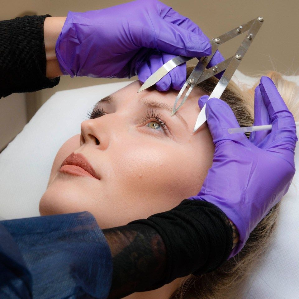 Mani guantate che utilizzano uno strumento per modellare le sopracciglia su una donna sdraiata durante un trattamento di bellezza.
