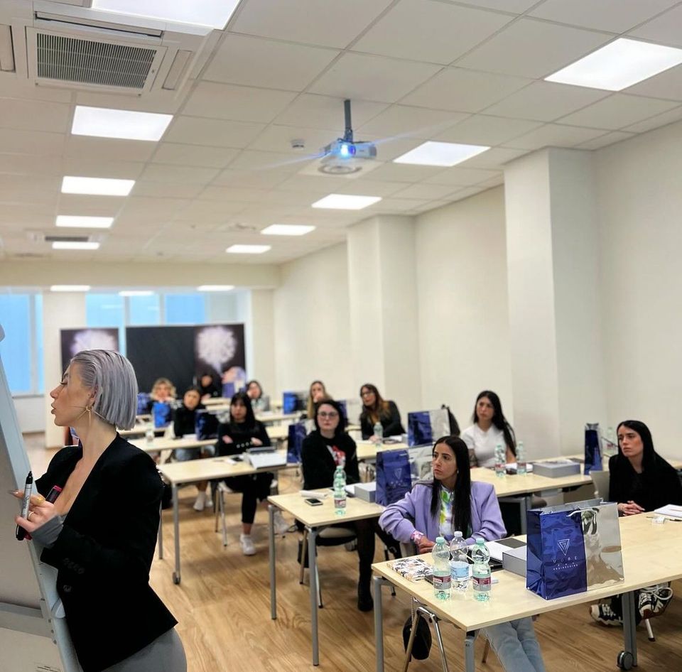 Sessione di formazione in aula con un relatore e partecipanti seduti ai banchi, illuminati da luci a soffitto.