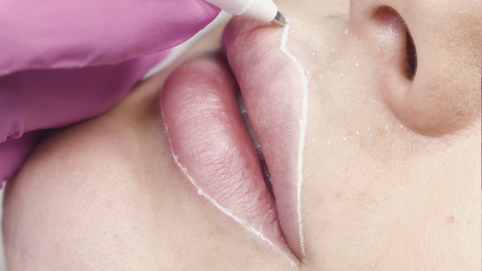 Primo piano di labbra e naso con un applicatore rosa che sfiora il contorno delle labbra.