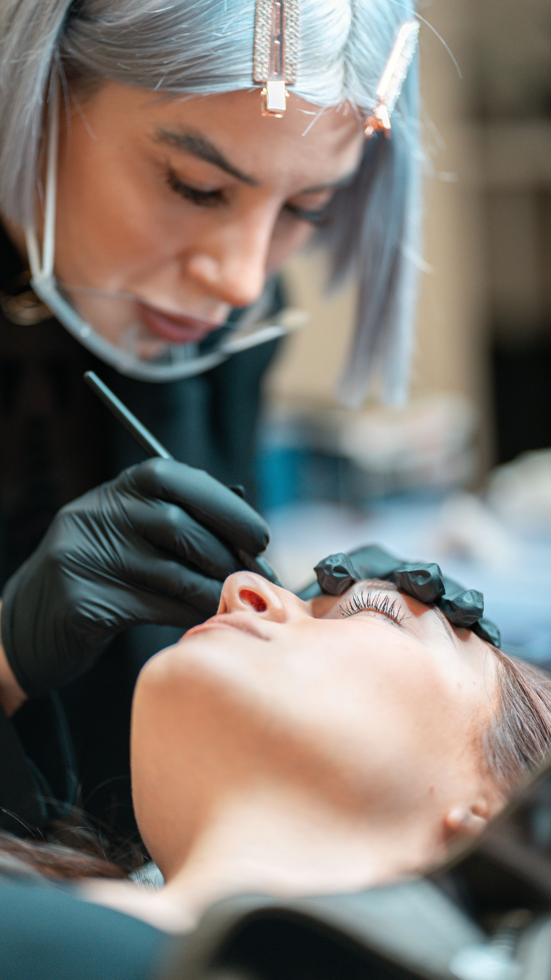 Una truccatrice applica il trucco agli occhi di una cliente sdraiata, con guanti neri e capelli argentati ben visibili.