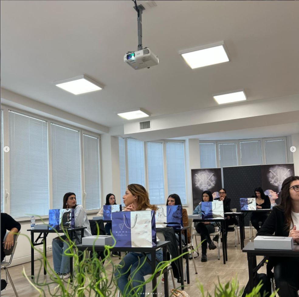 In un'aula luminosa, gli studenti siedono ai banchi e ascoltano una presentazione proiettata su uno schermo.