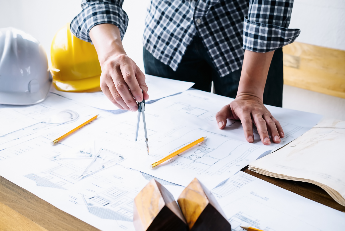 Architect working on blueprints, using compass, hard hats on table.