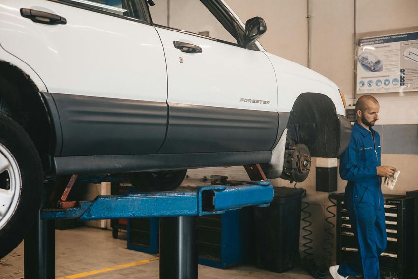 White SUV on a blue lift in an auto repair shop, with a mechanic standing beside it.