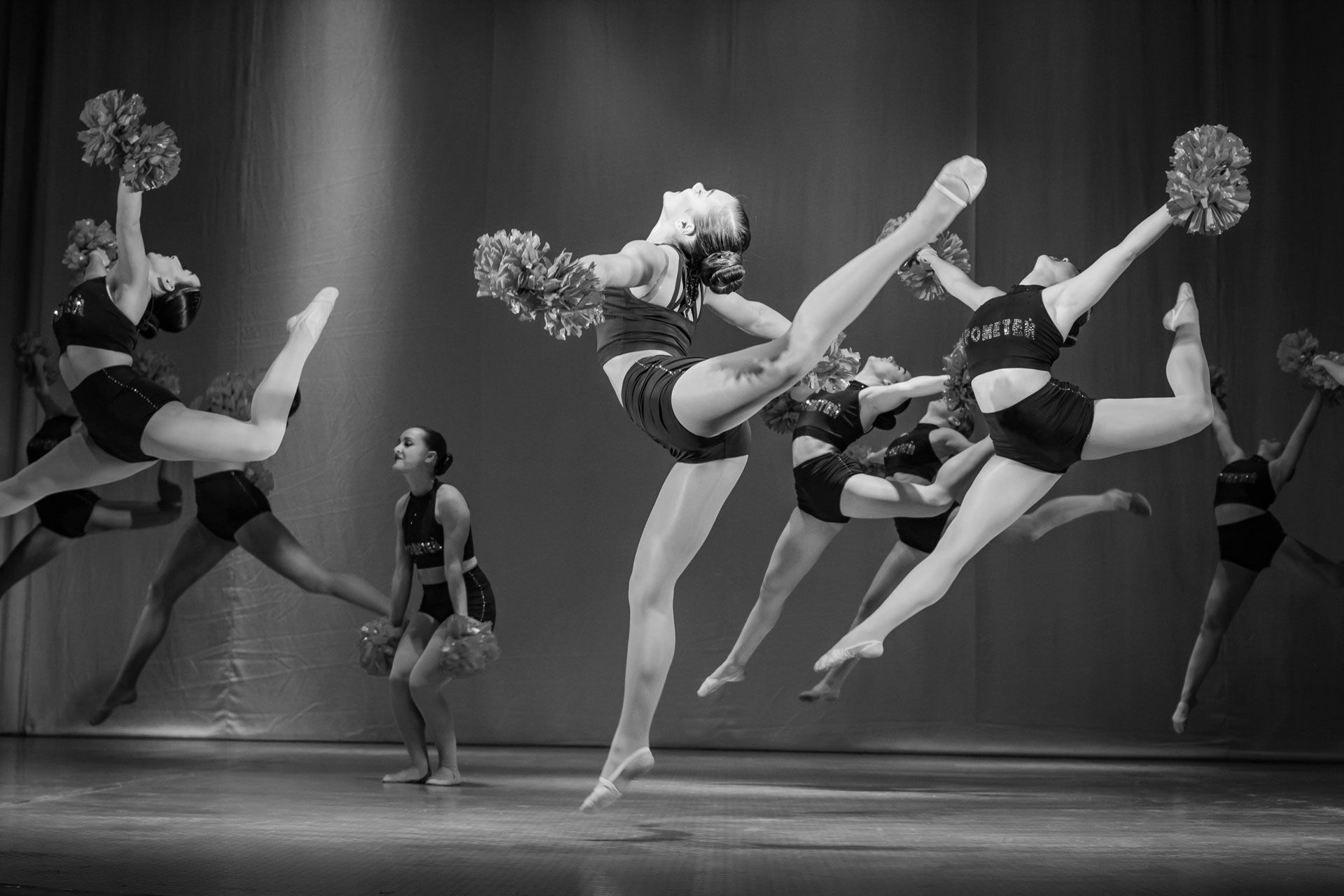 Cheerleaders mid-performance on stage, leaping and kicking with pom-poms in a black-and-white scene