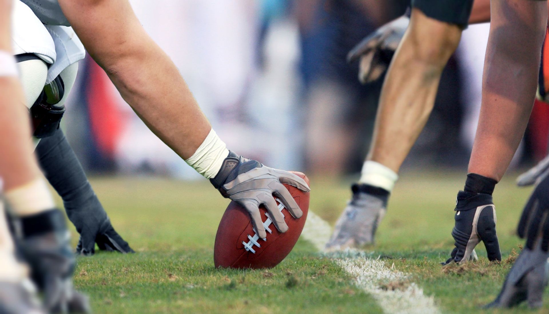 Football players line up at the line of scrimmage, one hand on the ball on a grassy field.