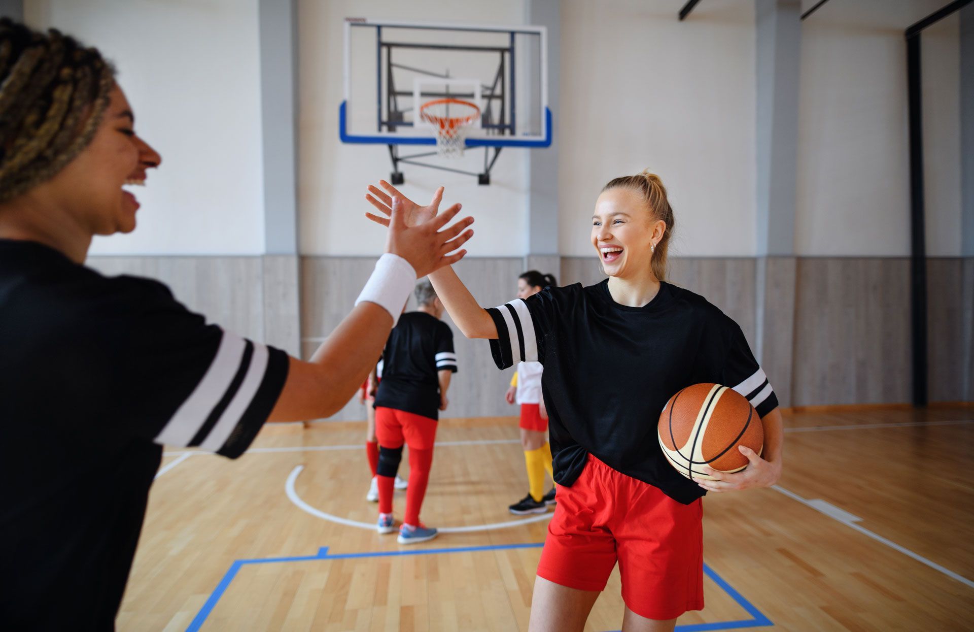 Two basketball players high-five on a gym court, one holding a ball near the hoop.