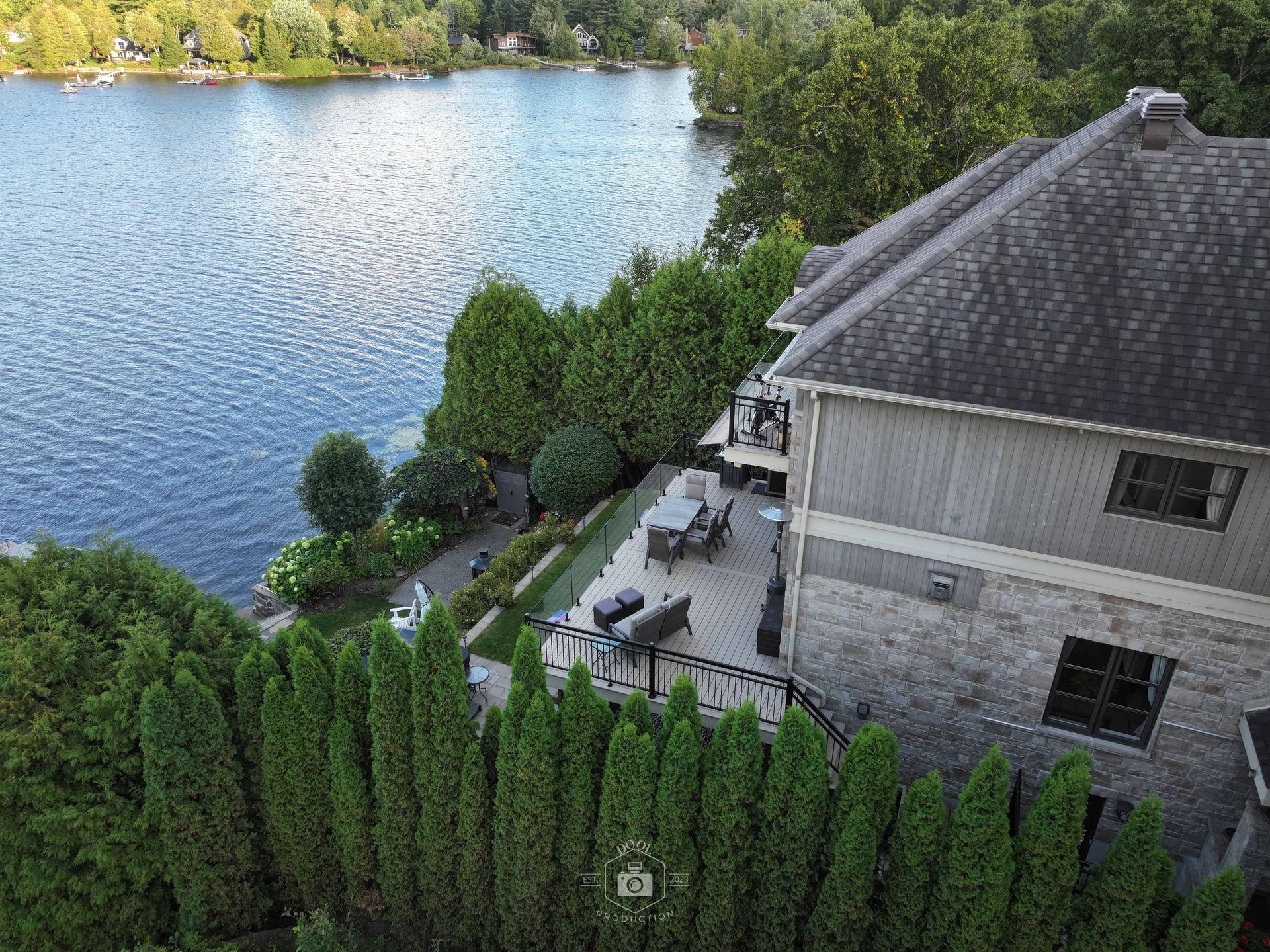 Maison avec façade en pierre et terrasse donnant sur un lac bleu, entourée d'arbres verdoyants.