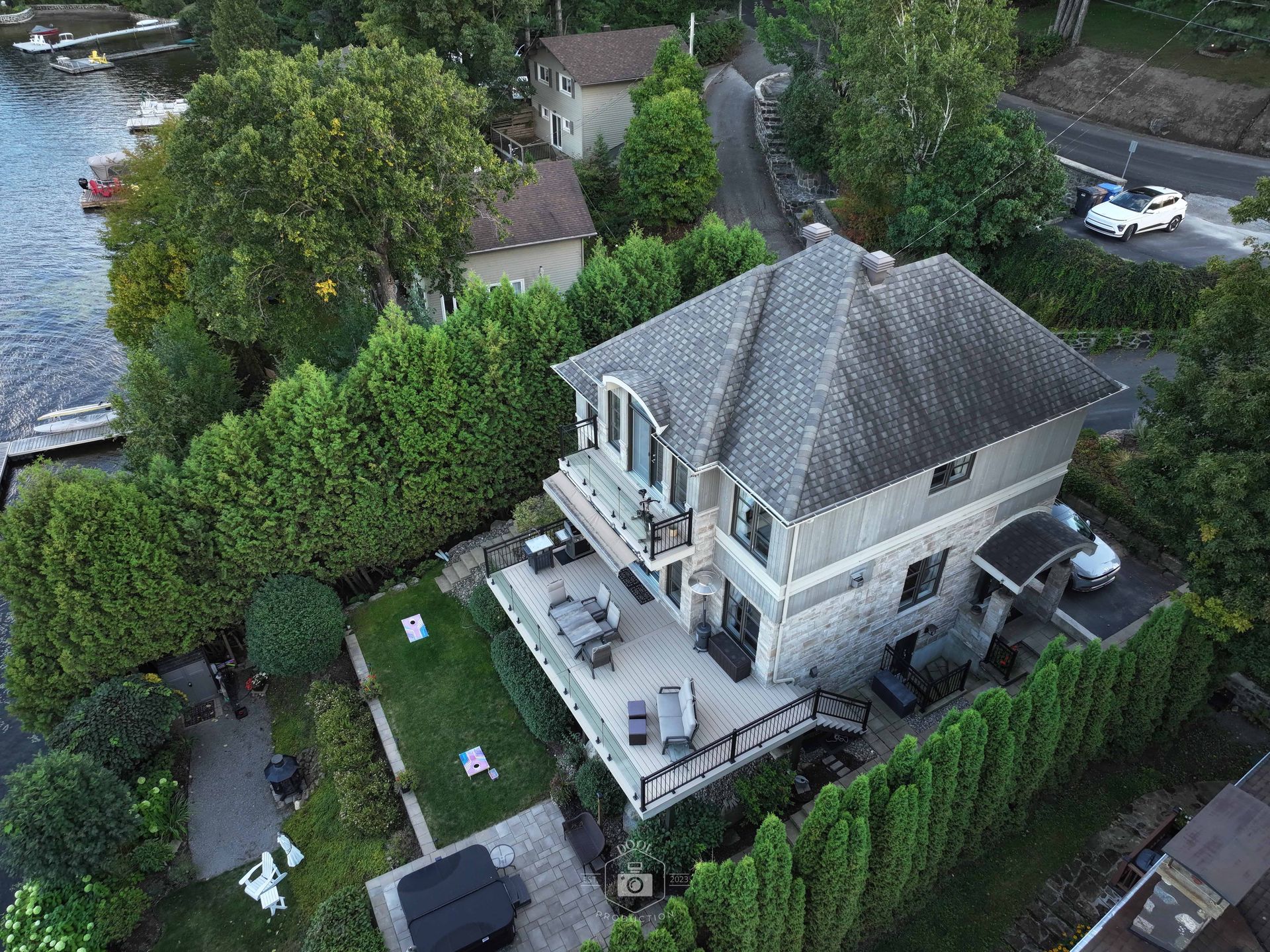 Vue aérienne d'une maison grise à deux étages avec une terrasse en bois, entourée d'arbres verts et donnant sur un lac.